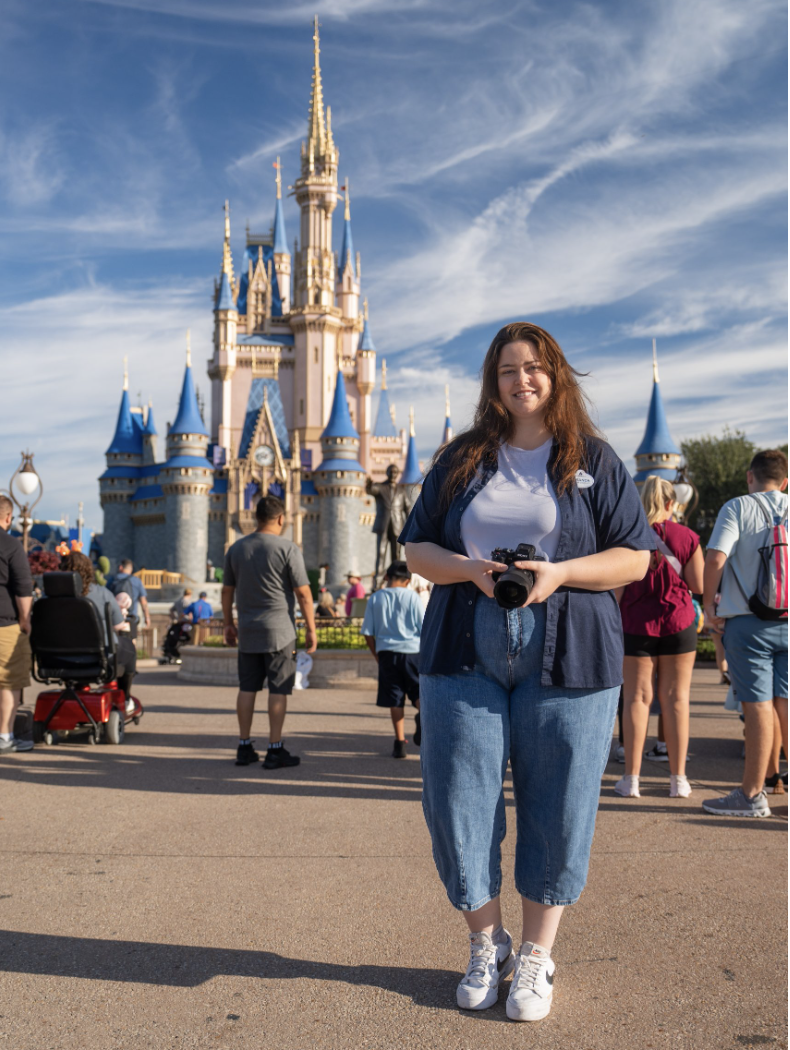 Person standing with a camera in front of a castle during the day, surrounded by visitors.