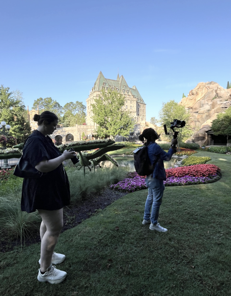 Two people photographing in a landscaped garden with flower beds and a large stone building behind them.