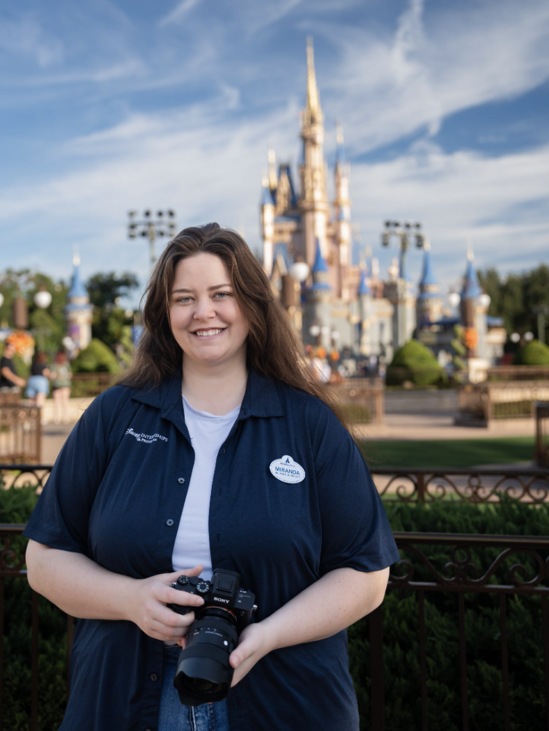 Person holding a camera in front of a castle during the day, wearing a dark shirt with a name tag.
