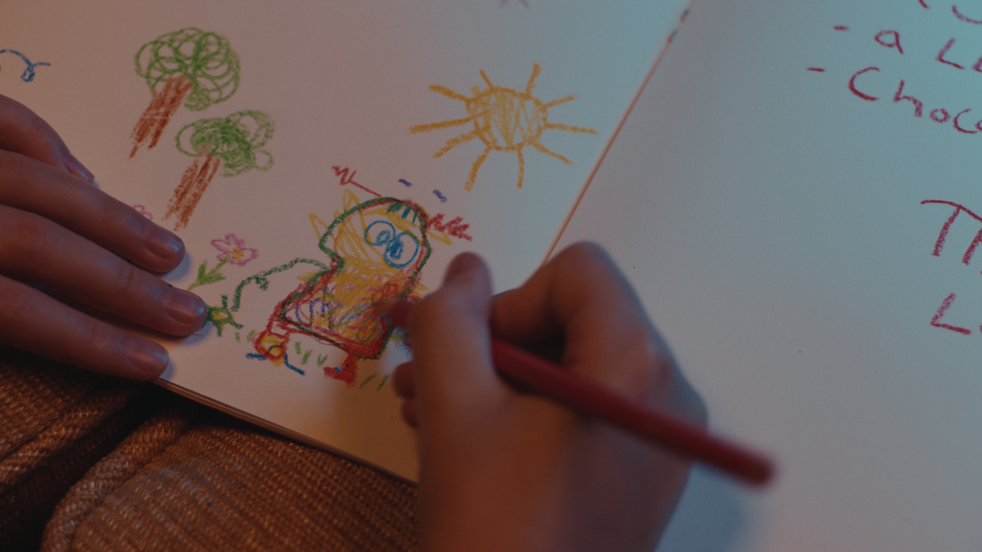 Close-up of a child&rsquo;s hands drawing a colorful character in a sketchbook with crayons, surrounded by doodles of trees, sun, and handwritten notes.