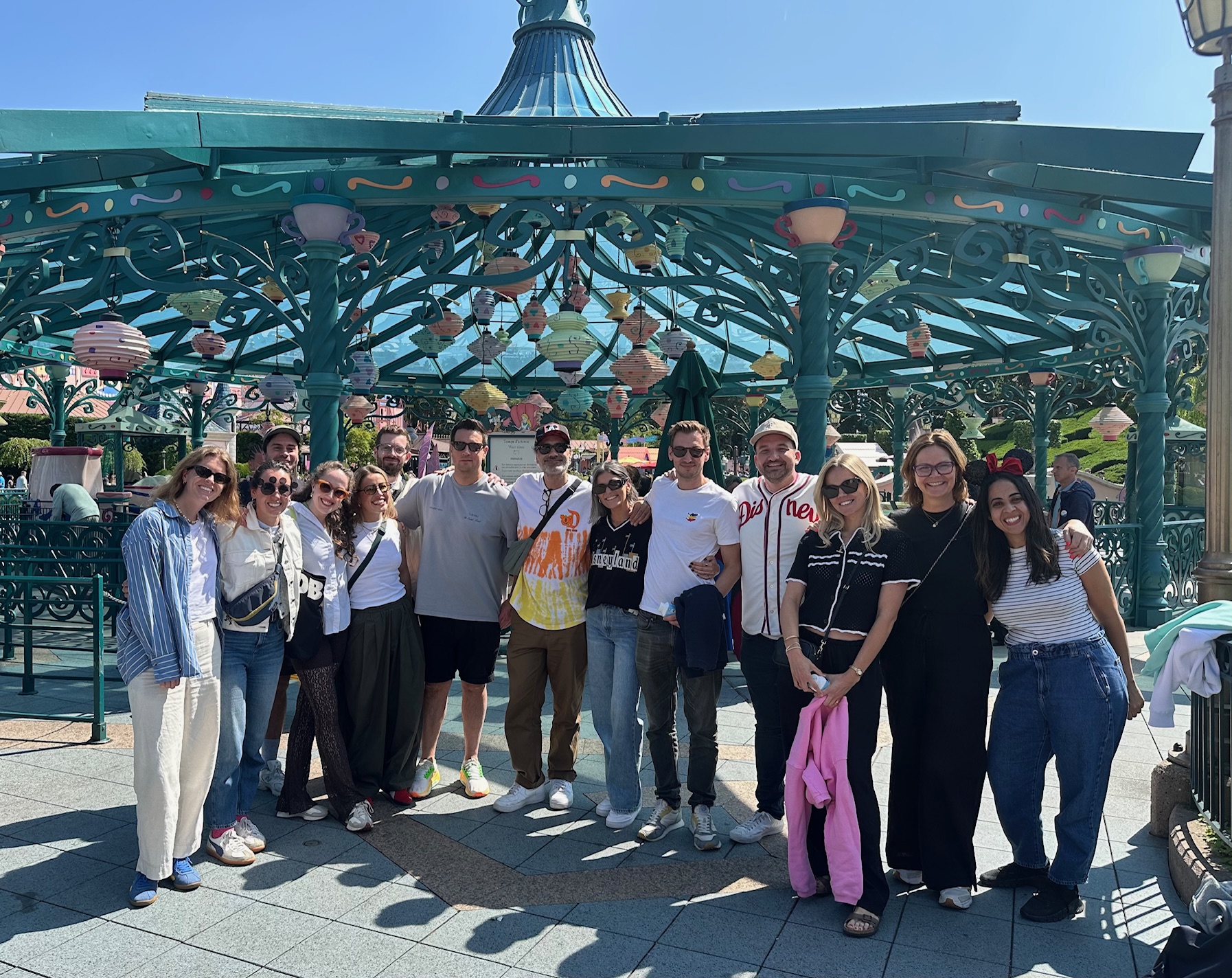 A group of people posing under the ornate teal Mad Hatter&rsquo;s Tea Cups pavilion at Disneyland Paris, decorated with colorful hanging teacups and surrounded by bright sunlight and greenery.