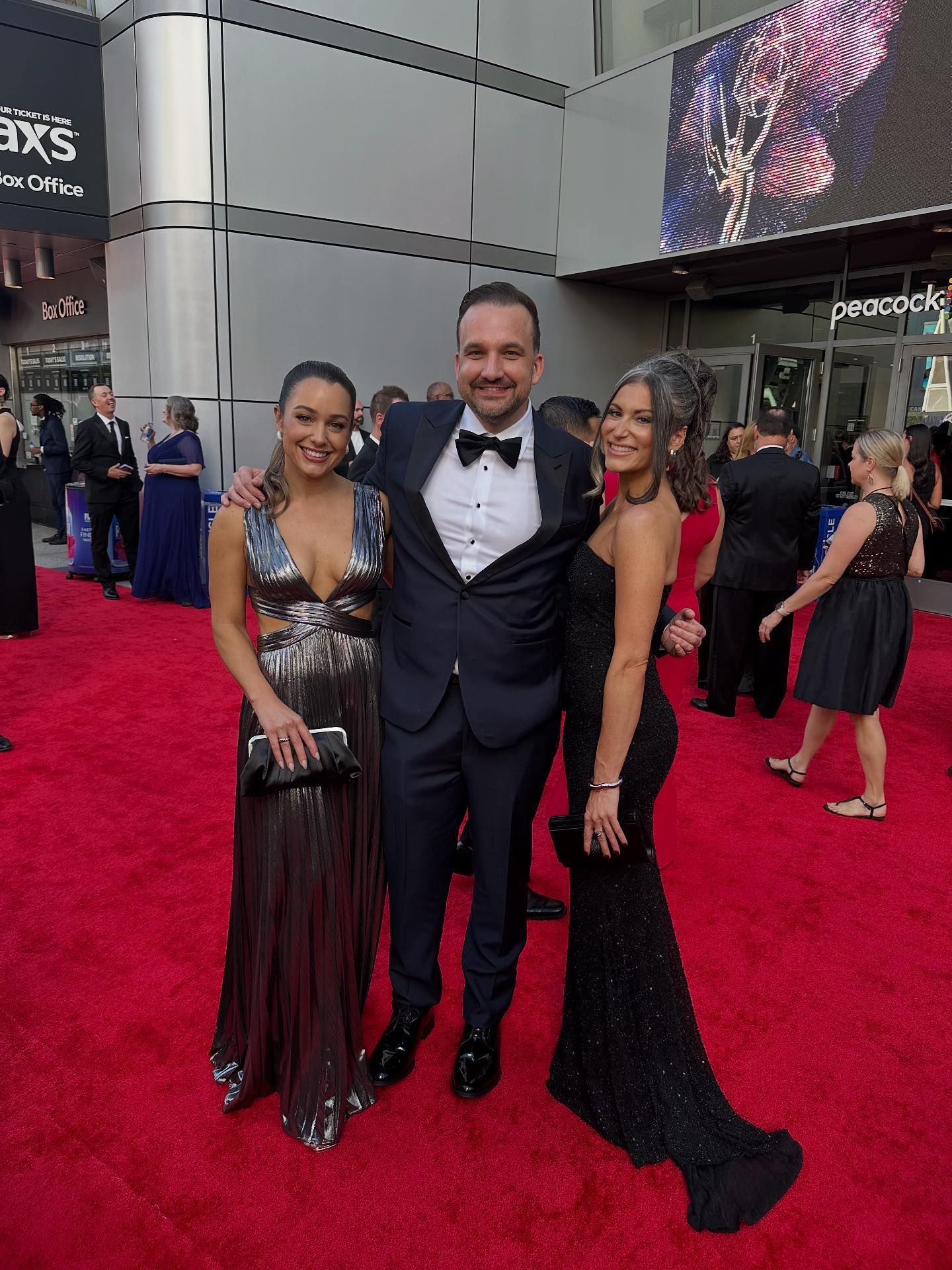 Three people dressed in formal evening attire posing on a red carpet at an awards event, with other attendees and a large screen visible in the background.