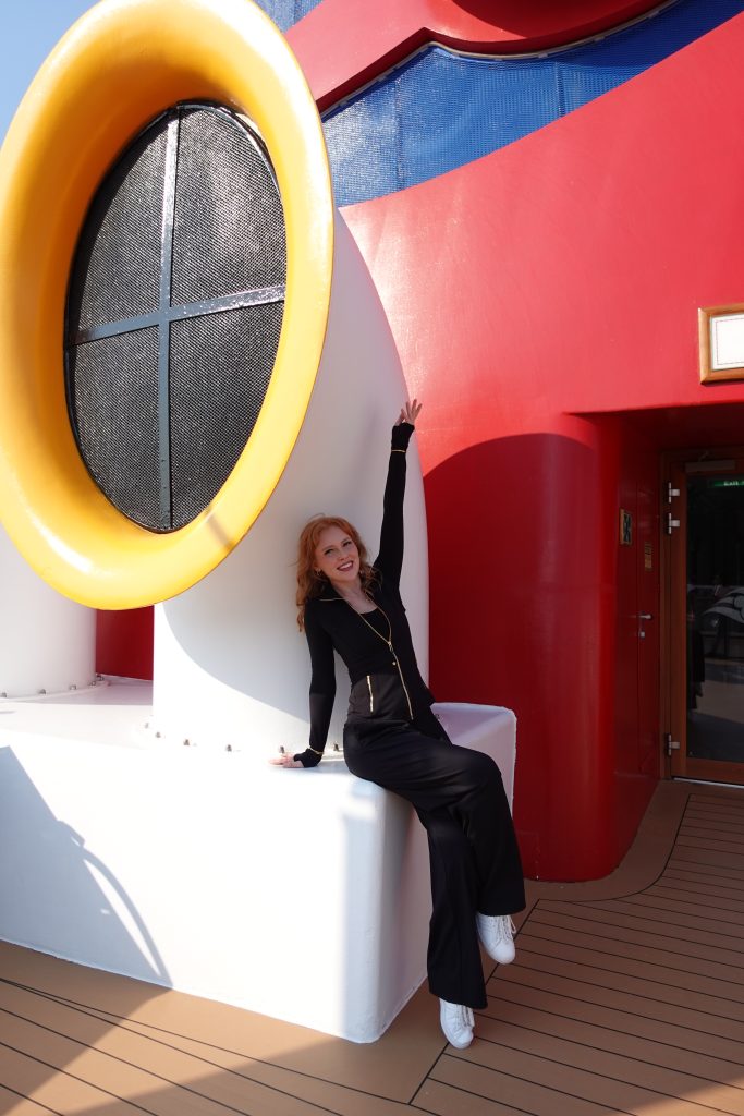 Image of Caroline, Performer sitting in front of Disney Cruise Line funnel with arm raised and smiling