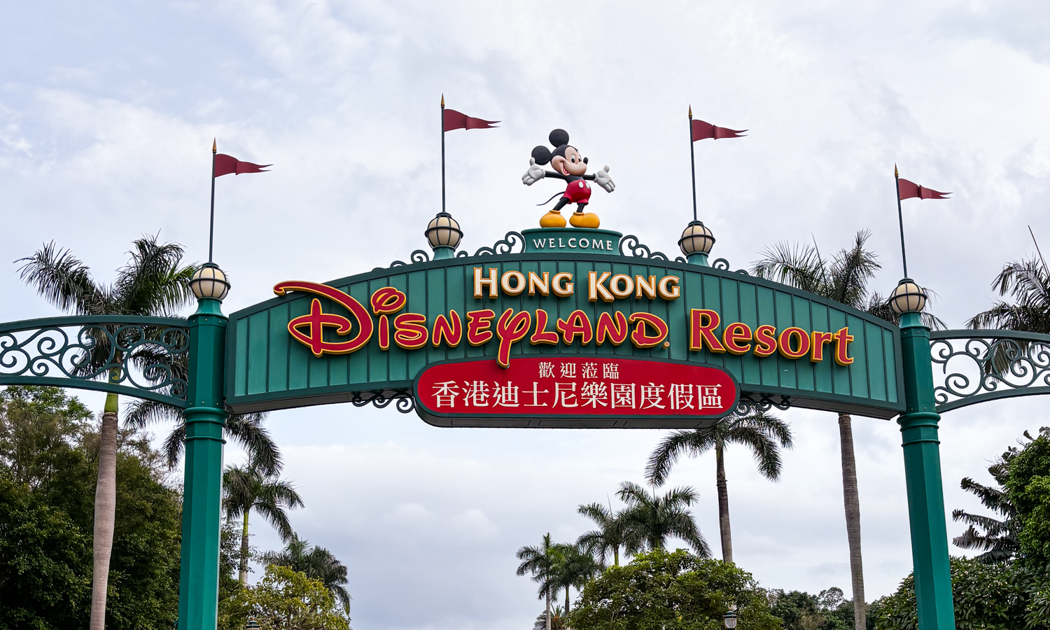 Hong Kong Disneyland Resort entrance signage in front of a cloudy sky and palm trees