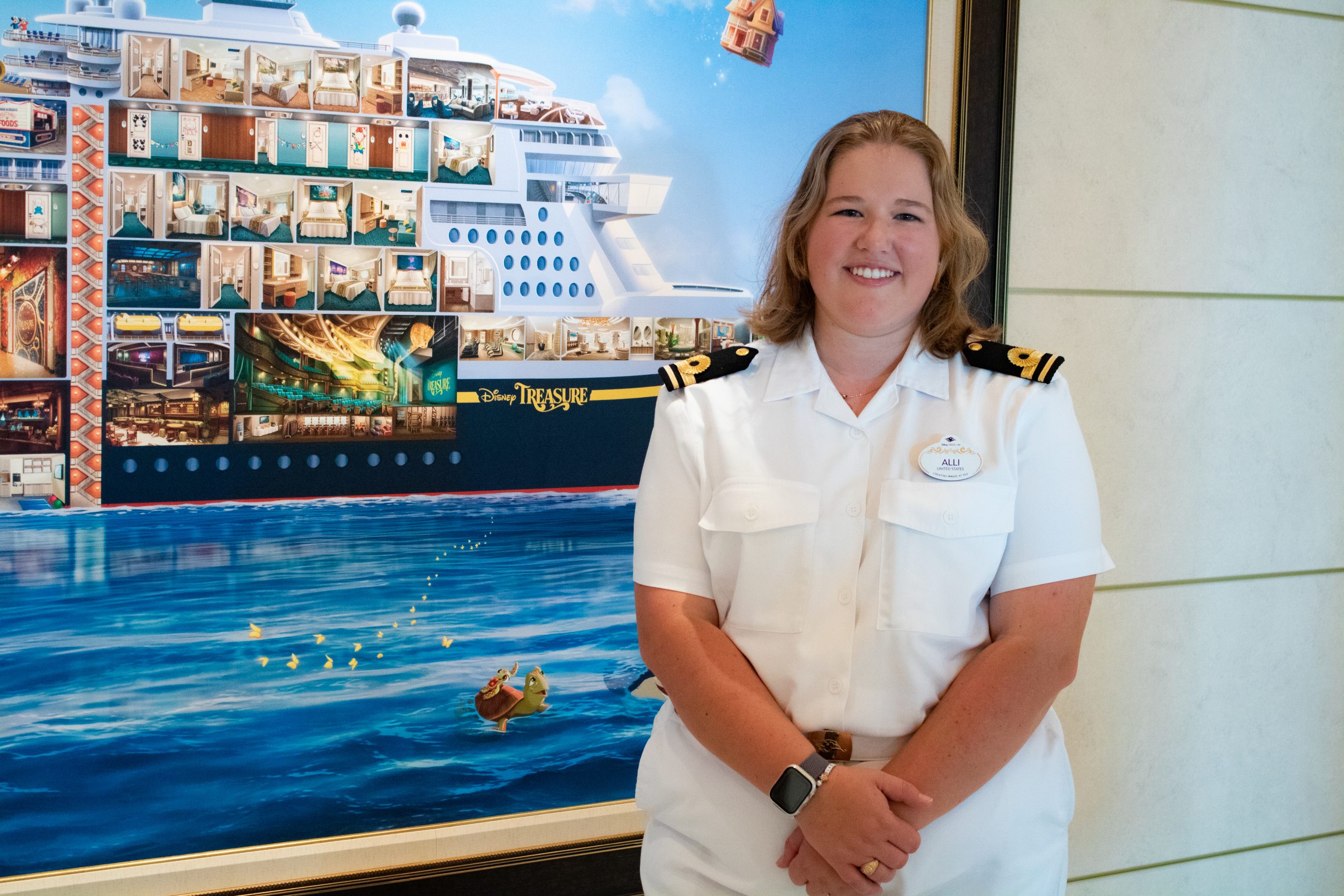 A crew member in uniform stands in front of a large decorative artwork or mural inside a cruise ship interior space.