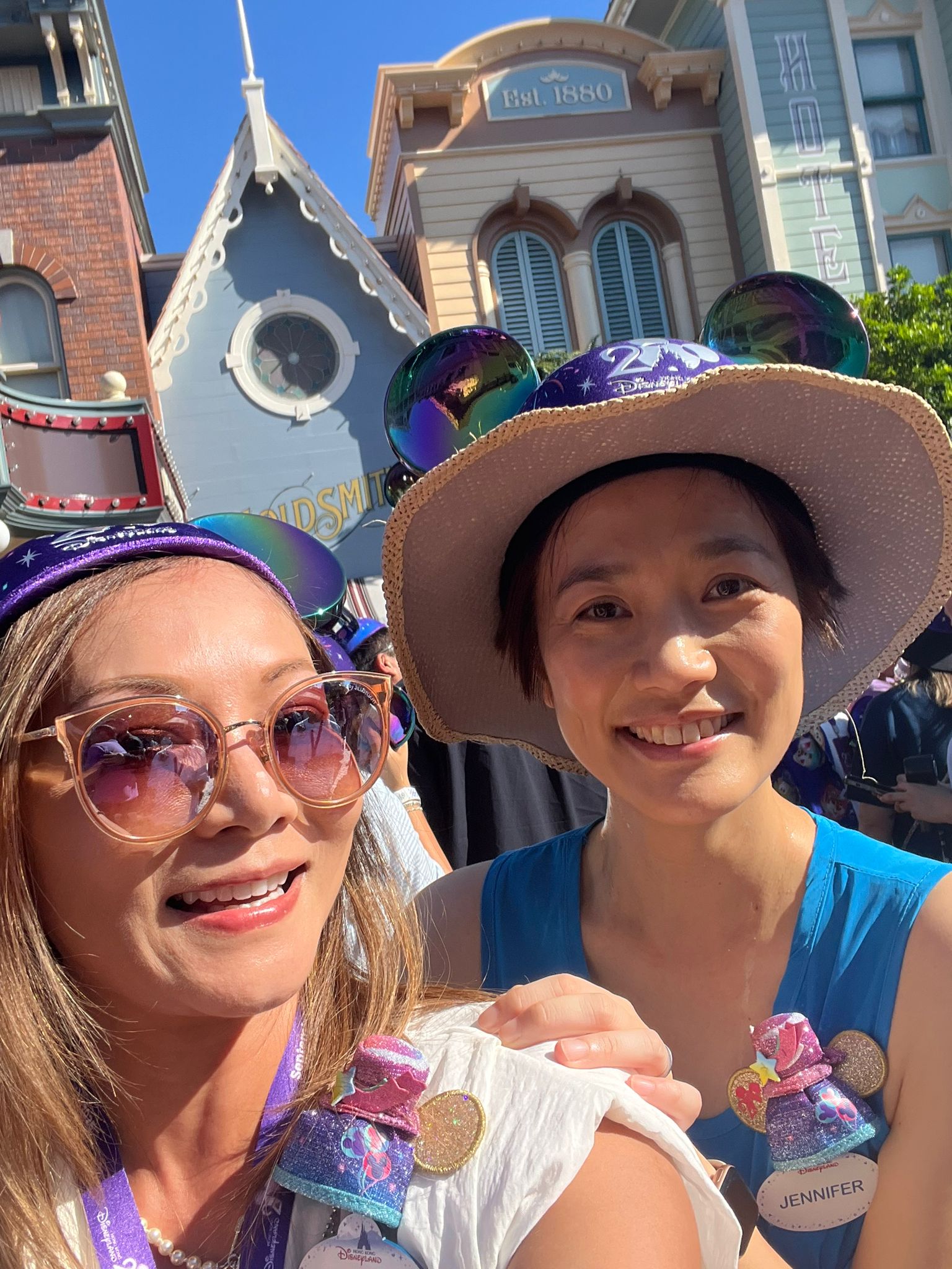 Close-up selfie of two people wearing themed hats with Mickey Mouse ears and decorative badges, standing in a crowded outdoor setting. Behind them are colorful, Victorian-style buildings.