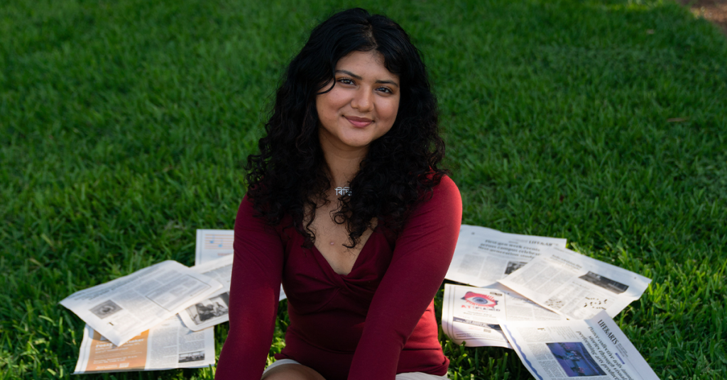 Person sitting on green grass wearing a long-sleeve red top and light-colored shorts, with several newspapers spread out around them, facing the camera in an outdoor setting.