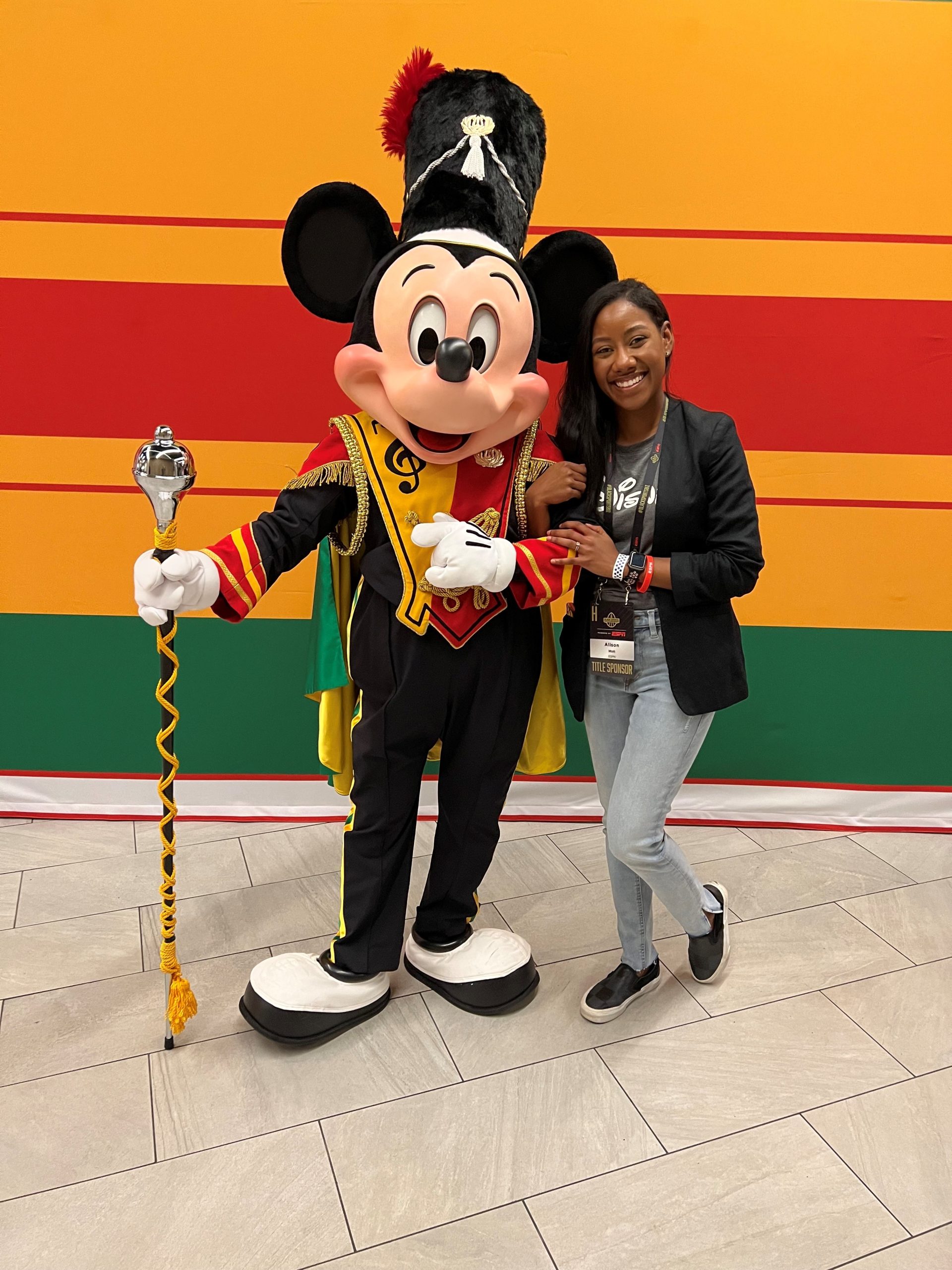 Photo of employee posing with Drum Major Mickey Mouse in front of a yellow, red and green striped backdrop.