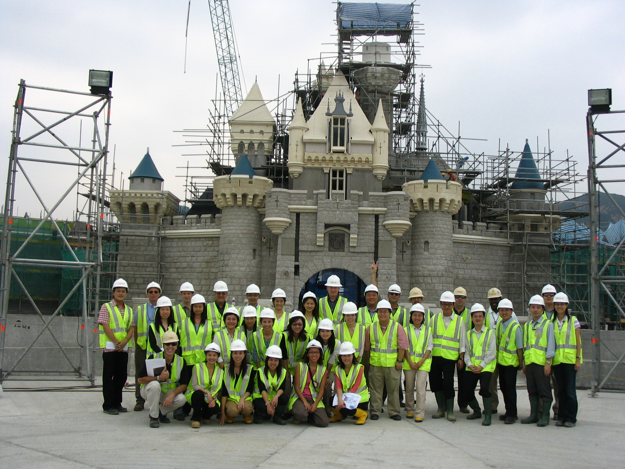 A large group of people wearing white hard hats and high-visibility safety vests pose for a group photo in front of a castle under construction. The structure features stone walls and turreted towers, surrounded by scaffolding and construction equipment, with a crane visible in the background.