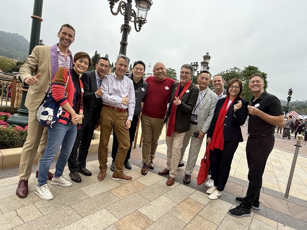 A group of about ten people stand together on a paved plaza outdoors, posing for a photo. Several wear lanyards and casual or business-casual clothing, with some giving thumbs-up gestures. Decorative lampposts, landscaped greenery, and distant hills or mountains appear in the background under an overcast sky.