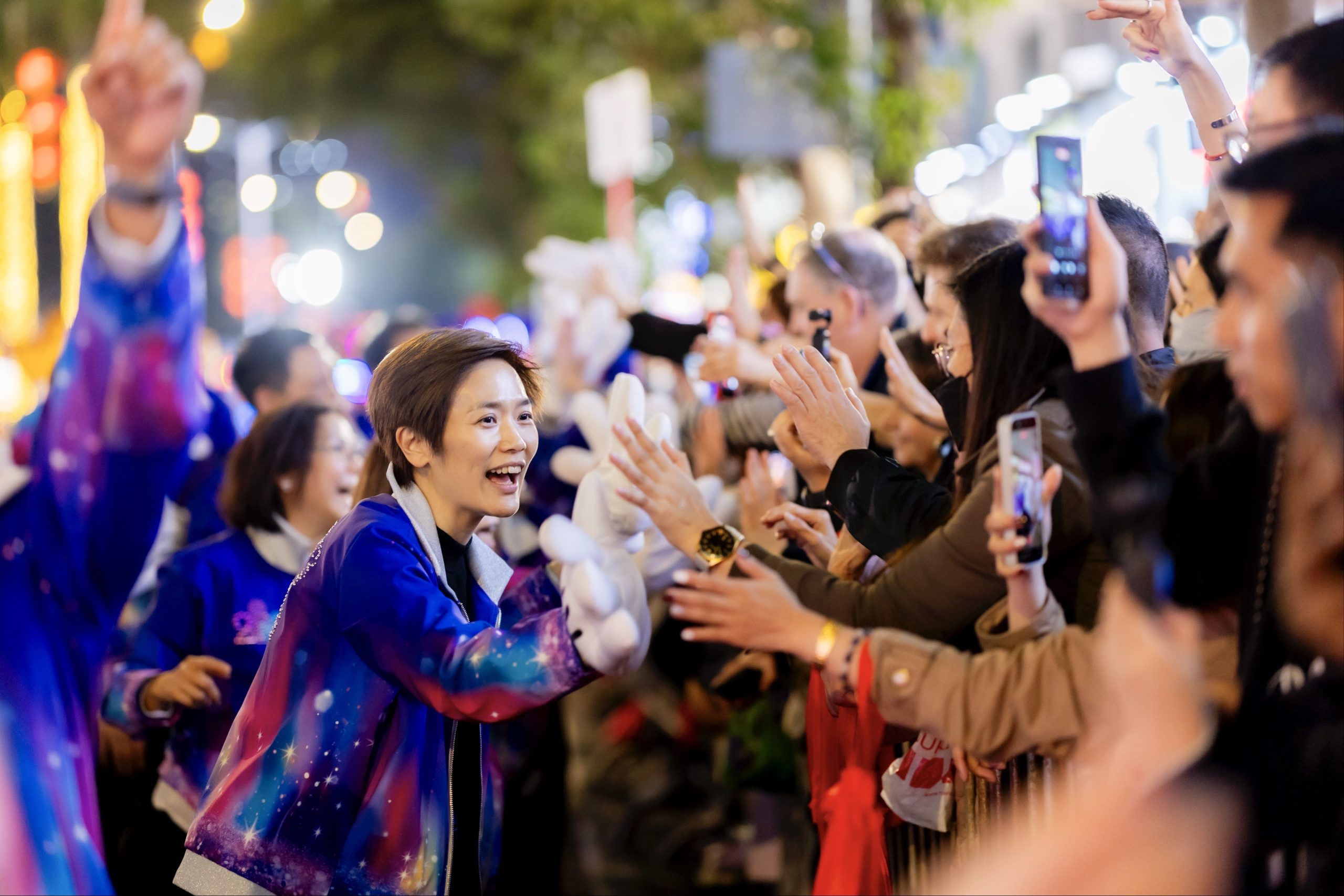 A person in a colorful jacket with a galaxy pattern reaches out to greet a crowd gathered along a street at night. Many people stand behind a barrier, extending their hands and holding up smartphones to take photos, while bright city lights and blurred signage glow in the background.