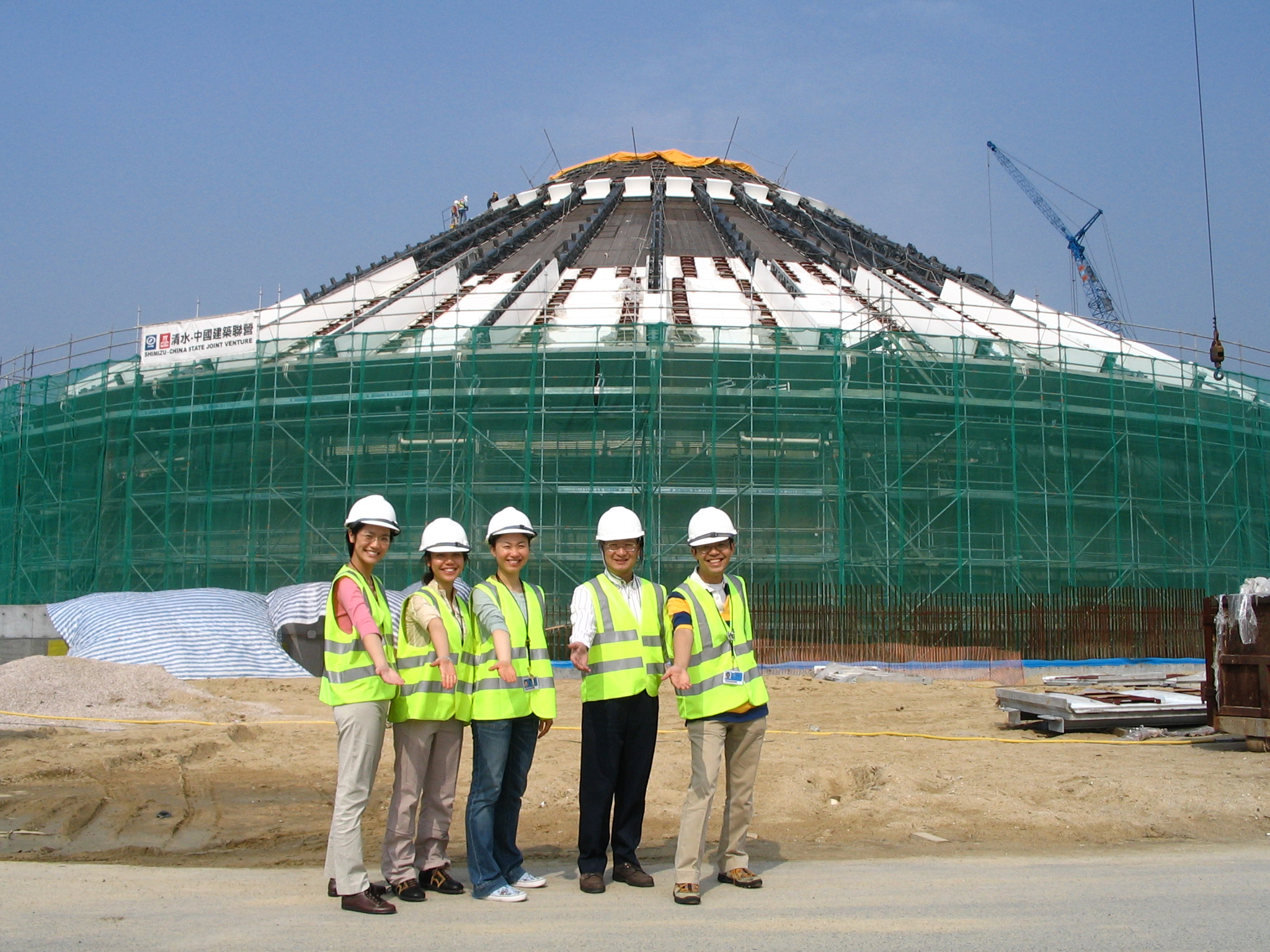 Five people wearing white hard hats and high-visibility safety vests stand in front of a large, dome-shaped building under construction. The structure is covered in green scaffolding and partially installed panels, with a crane visible nearby. The group stands on a dirt construction site with their arms extended forward, posing together in front of the project.