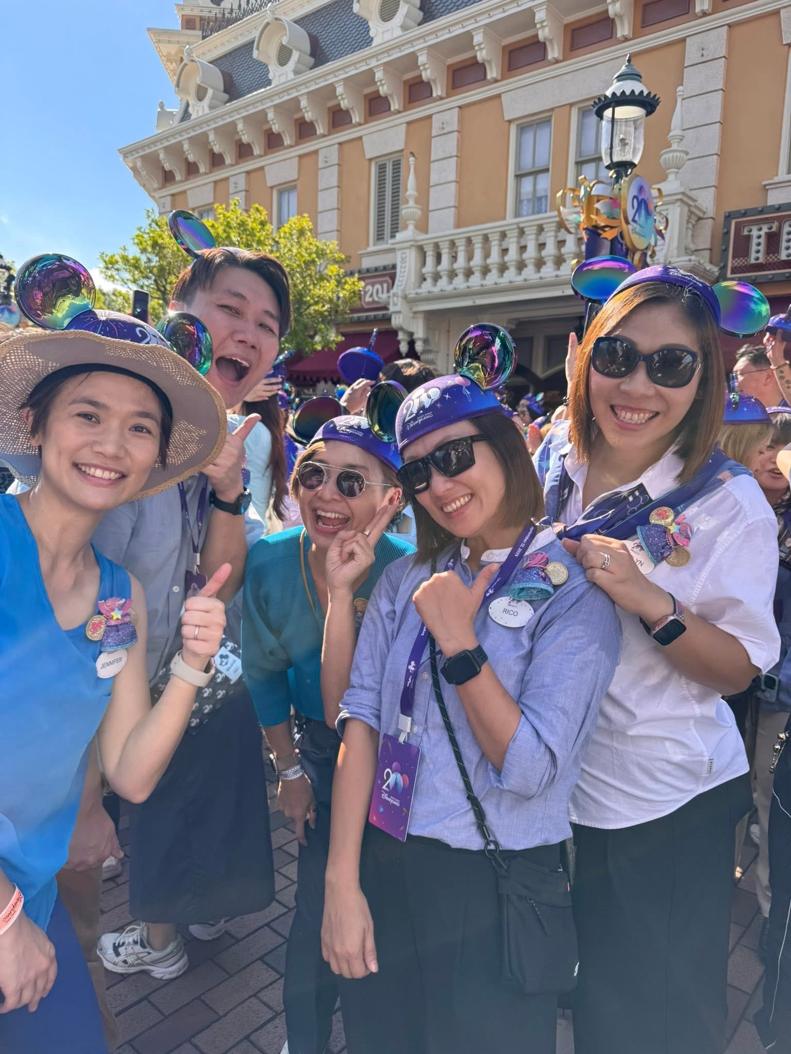 A group of five people pose together outdoors in a busy theme park setting, wearing matching Mickey Mouse ear hats and lanyards with badges. They stand in front of a decorative, historic-style building with ornate details and a lamppost featuring a &ldquo;20&rdquo; celebration sign, while other visitors and colorful structures fill the background under a clear blue sky.