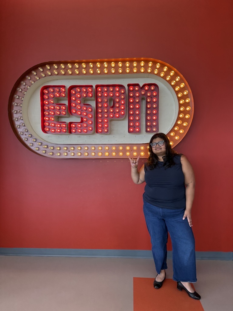 Person standing in front of an illuminated ESPN sign on a red wall, making a hand gesture while posing.