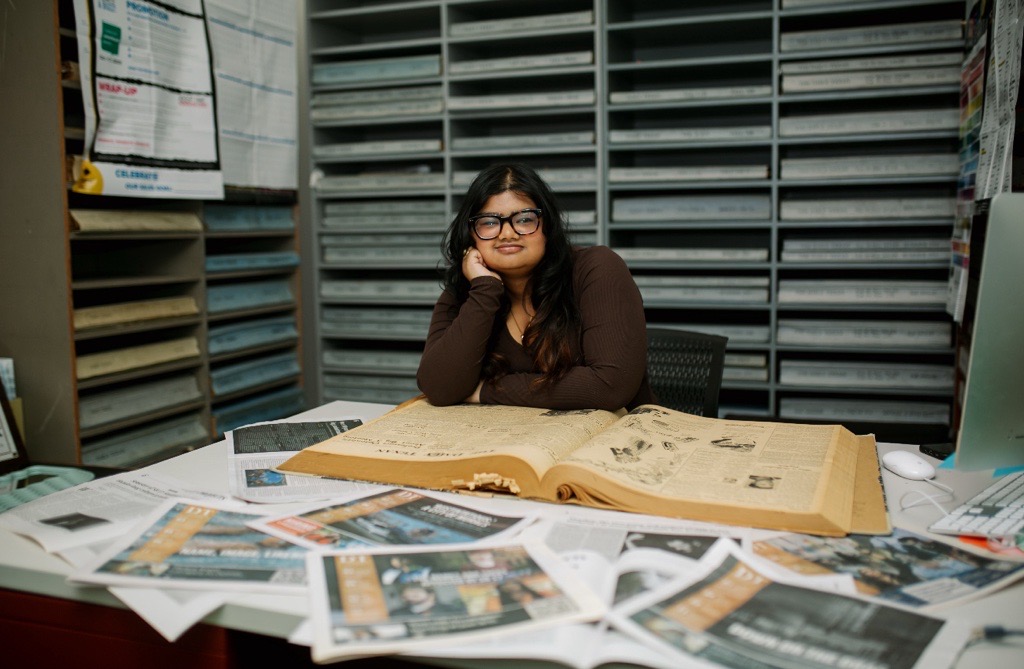 Person seated at a desk covered with newspapers and an open book, with organized shelves of archived materials in the background.