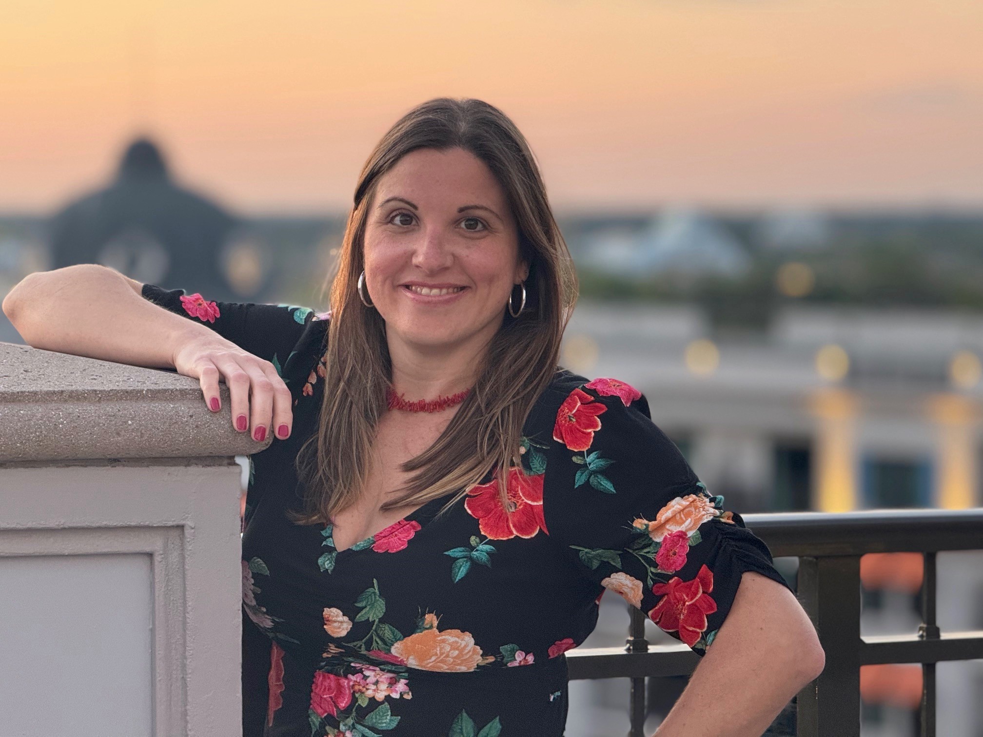 Photo of employee in a floral top with arm propped on a ledge in front of a sunset.