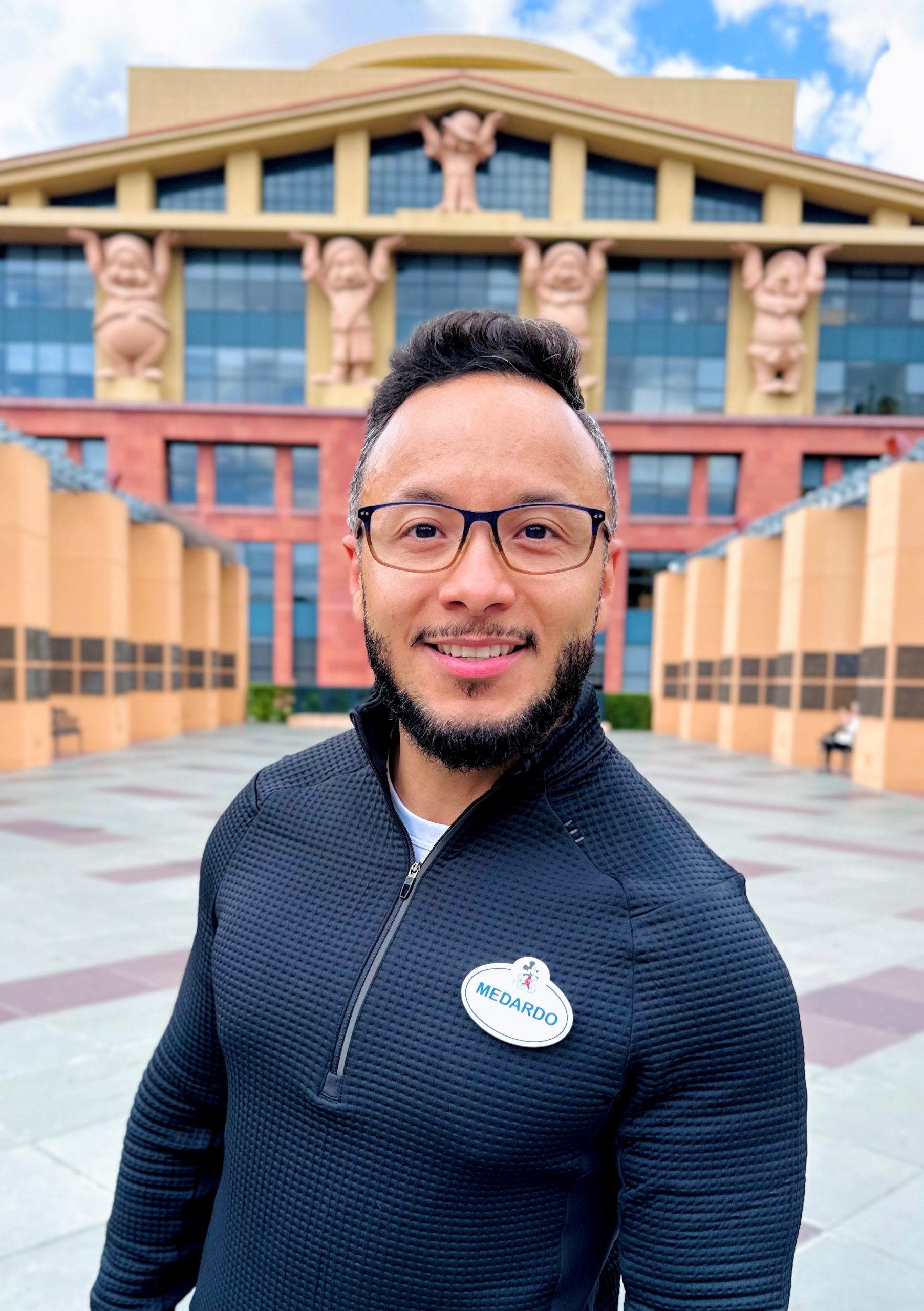 Smiling employee in a dark shirt in front of a Disney building.