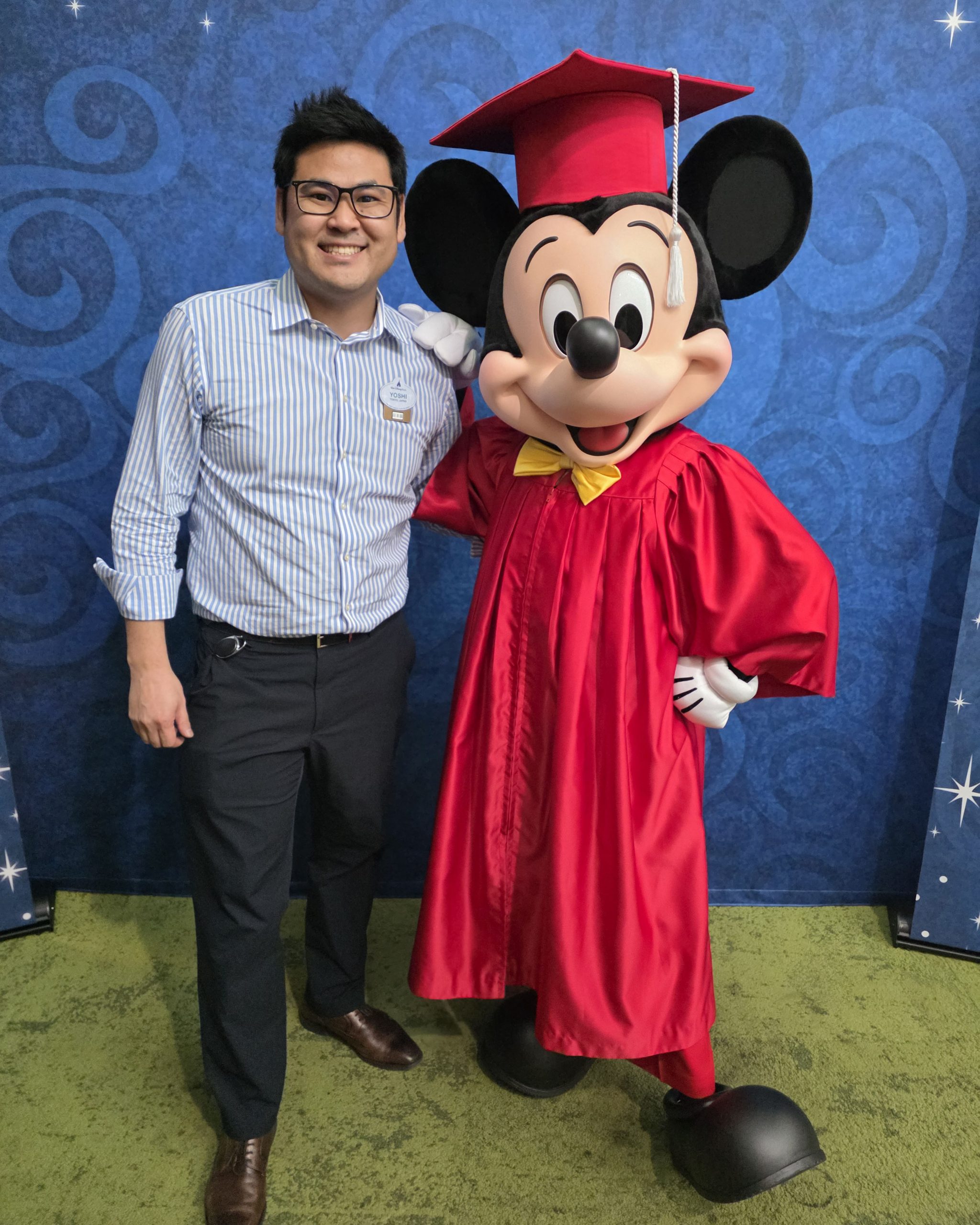 Disney recruiter Yoshi poses with Mickey Mouse in graduation attire, symbolizing his career growth from the Disney College Program to a leadership role in professional recruitment and talent acquisition.