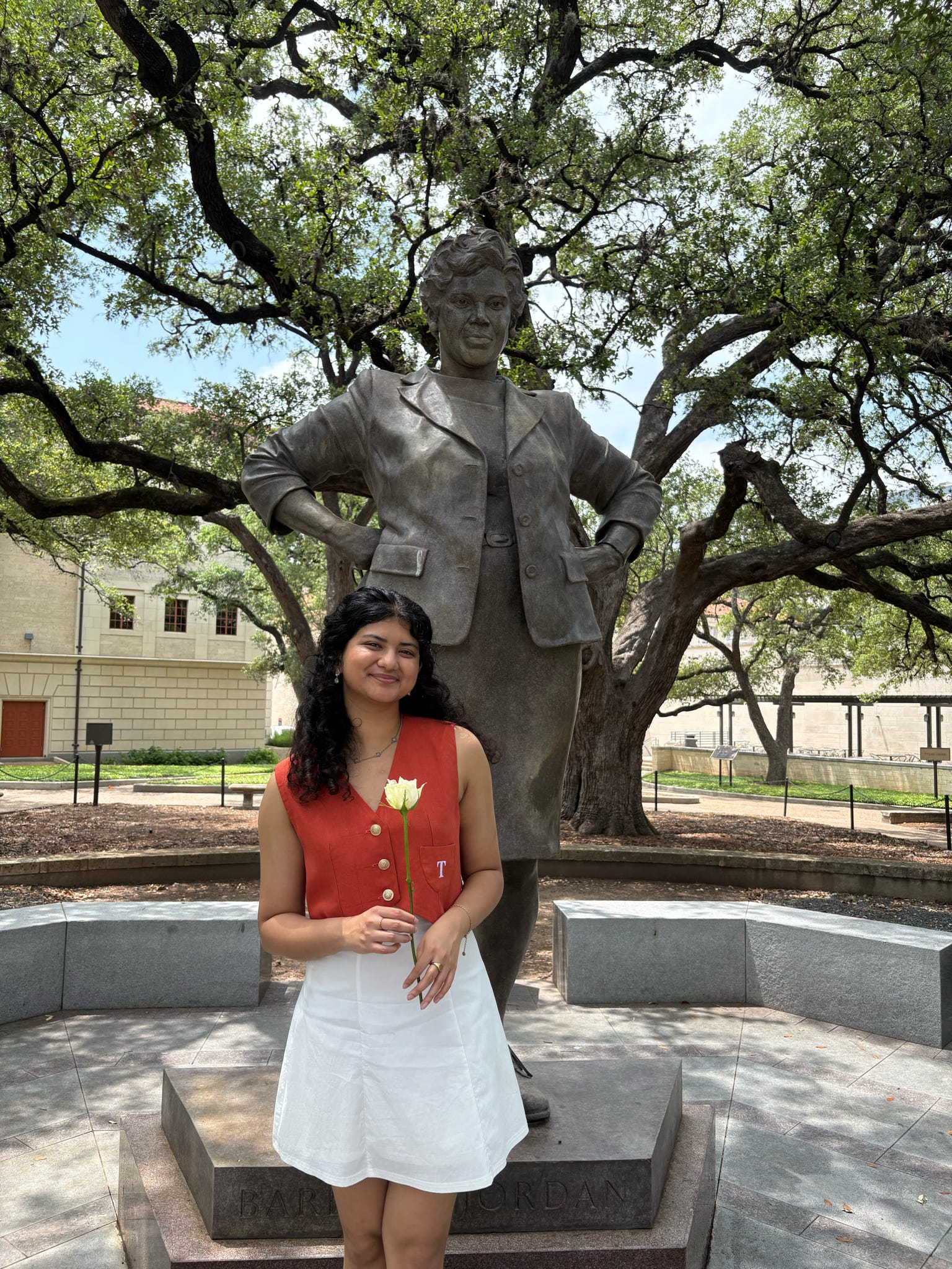 Person standing in front of a large bronze statue labeled &ldquo;Barbara Jordan&rdquo; in a landscaped campus setting, holding a white rose and wearing a reddish-orange sleeveless top with gold buttons and a white skirt; trees with spreading branches and light-colored buildings are visible in the background.
