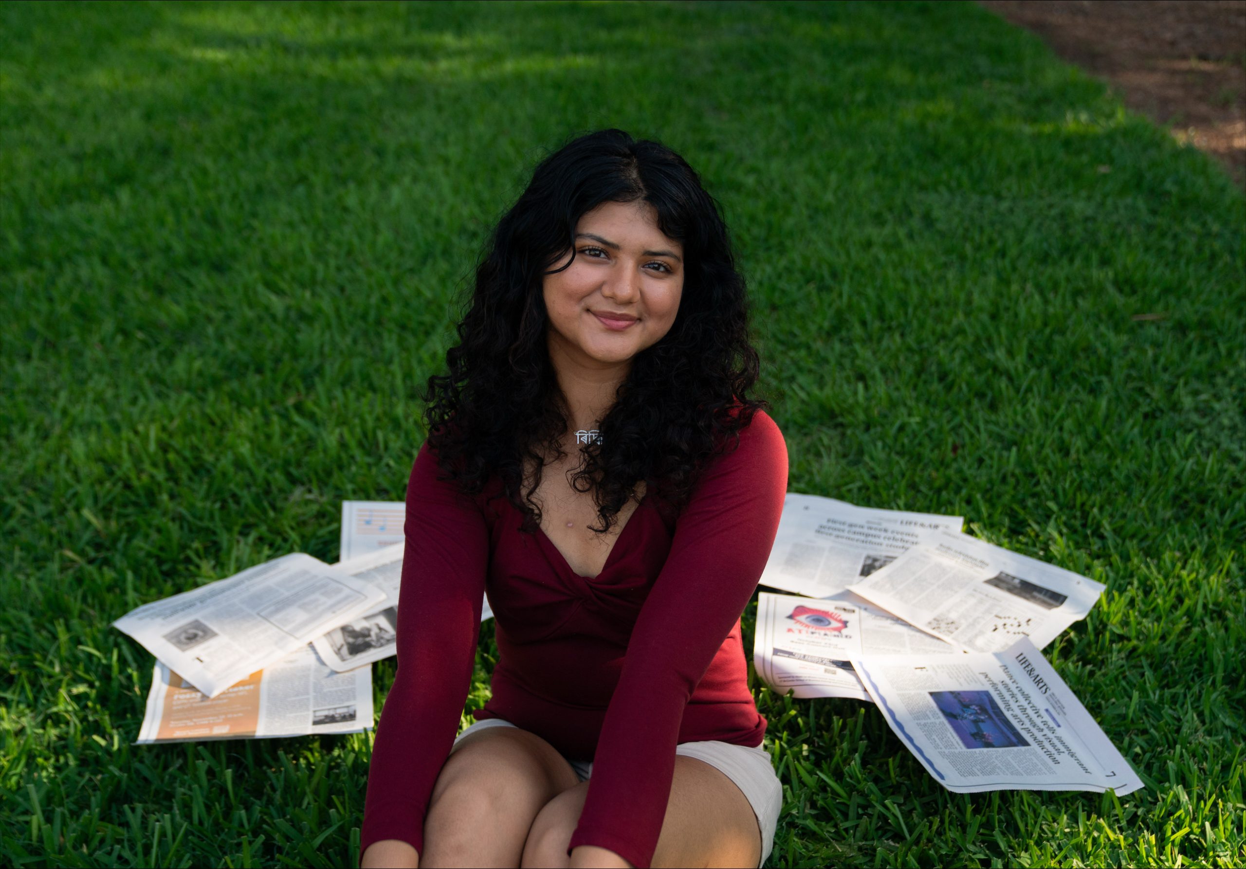 Person sitting on green grass wearing a long-sleeve red top and light-colored shorts, with several newspapers spread out around them, facing the camera in an outdoor setting.