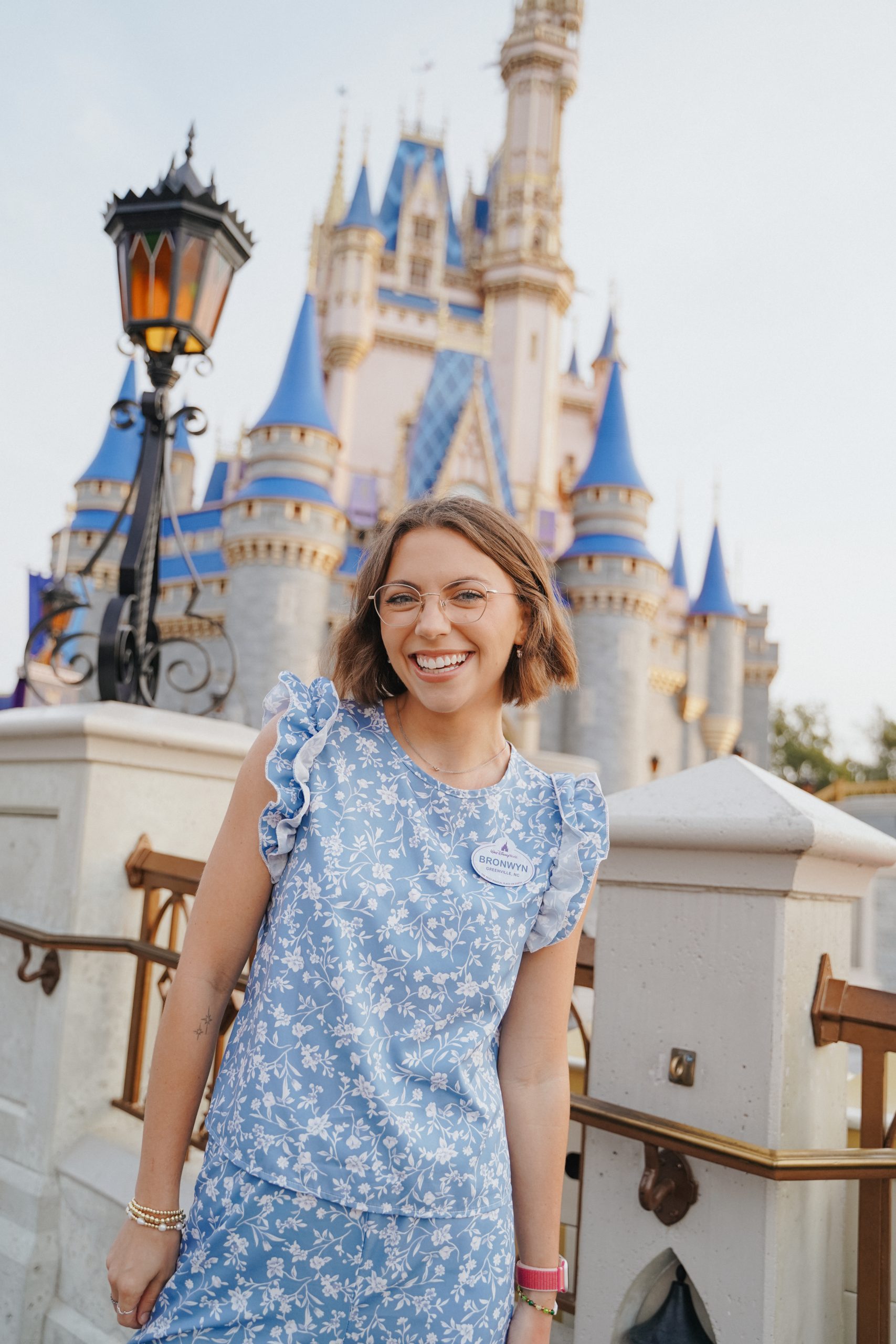 A woman wearing a light blue floral outfit and glasses stands on a bridge in front of a castle with blue spires, while a decorative lamp post and stone railings frame the scene in soft daylight.