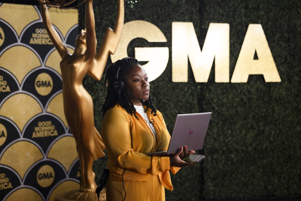Eboni, Senior Broadcast Producer for Good Morning America, works on a laptop inside the GMA studio environment. Good Morning America and GMA branding are visible behind her as she supports ABC News content production at Disney.