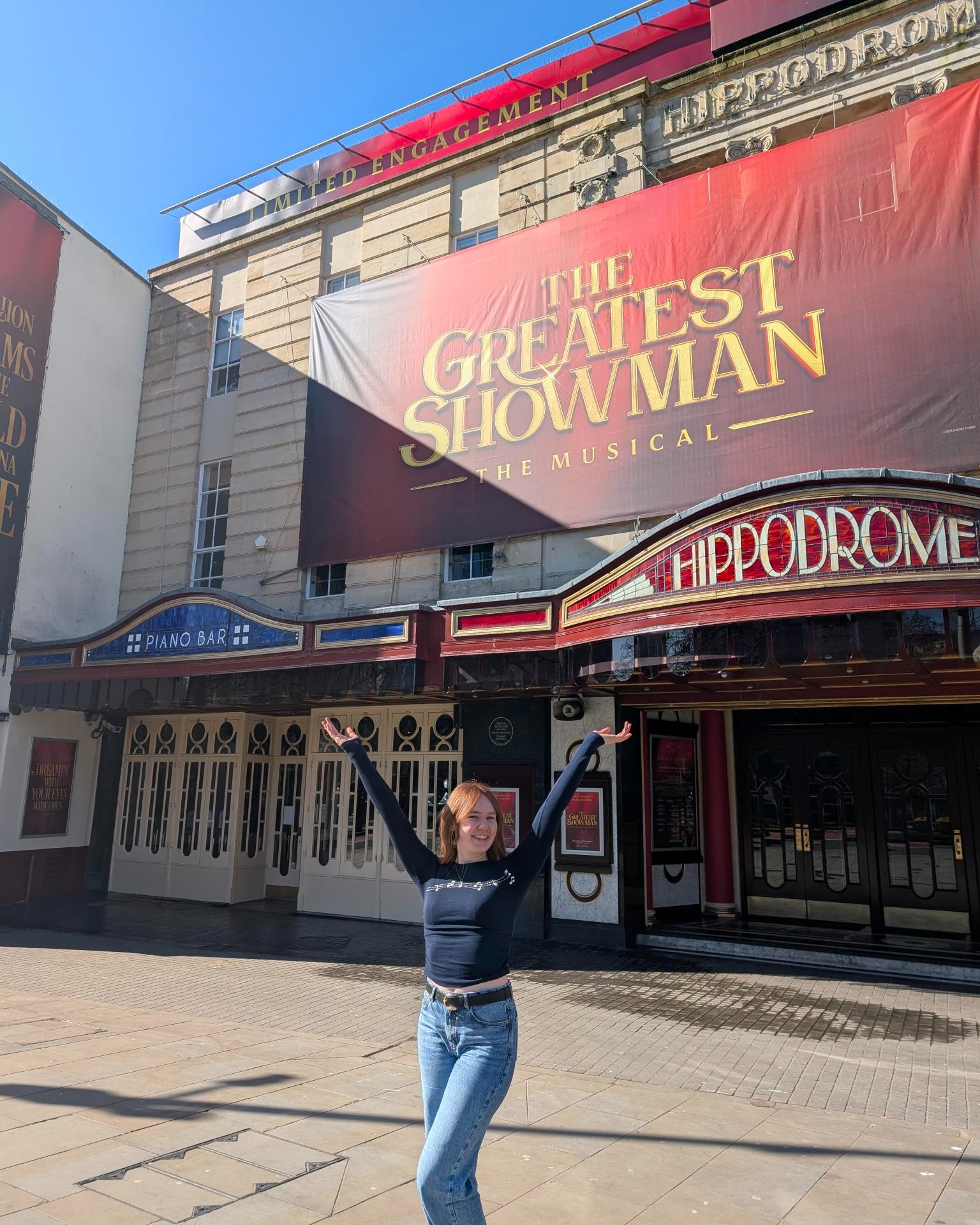 Disney EMEA Creative Venue Technician Apprentice Hannah smiling with her hands up while standing in front of a "The Greatest Showman" banner