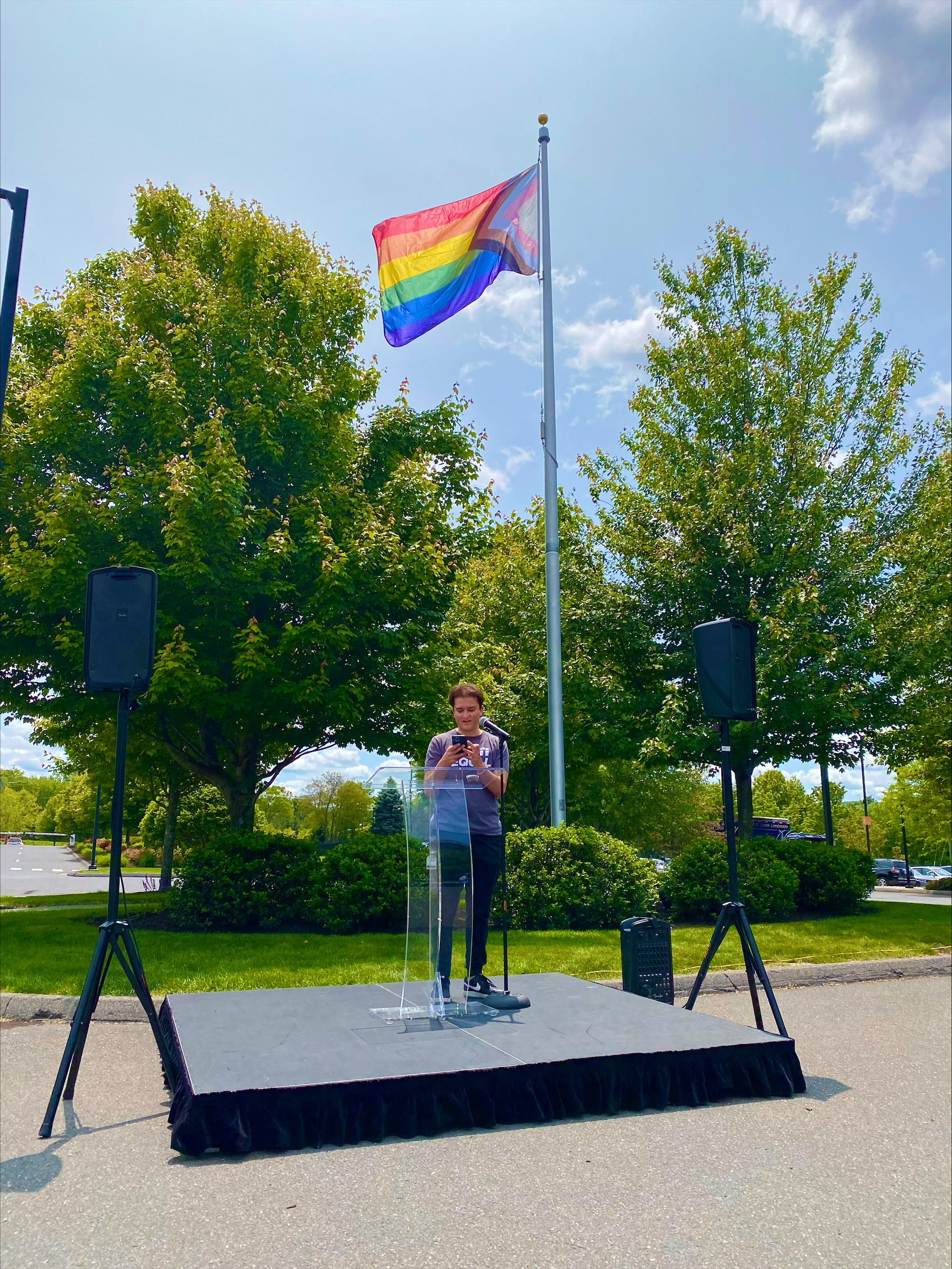 Mac speaks at an outdoor podium beneath a raised LGBTQ+ Pride flag, with trees and event speakers in the background.