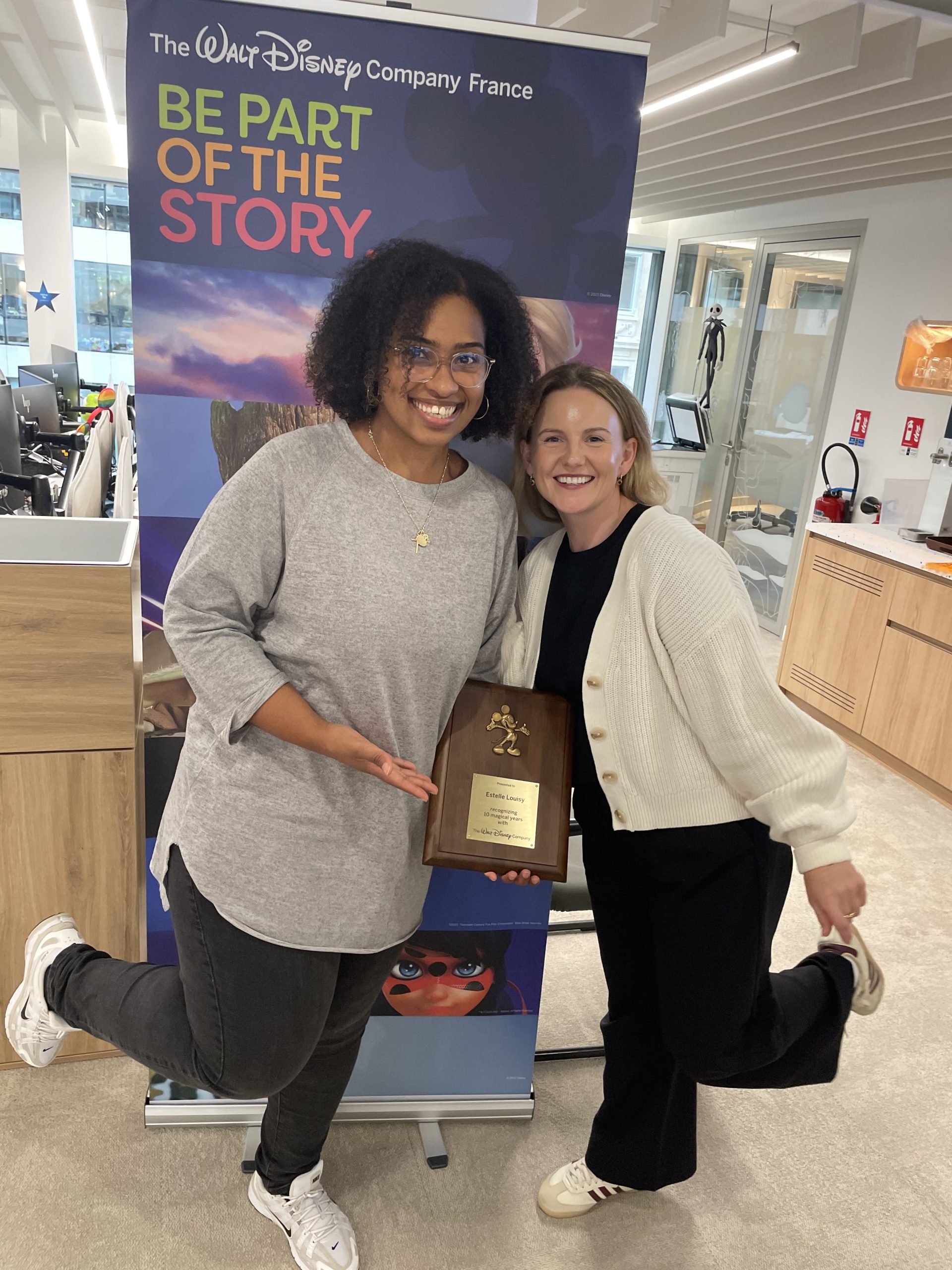 Two employees smile while holding an award beside signage reading &ldquo;The Walt Disney Company France&rdquo; and &ldquo;Be Part of the Story.&rdquo;