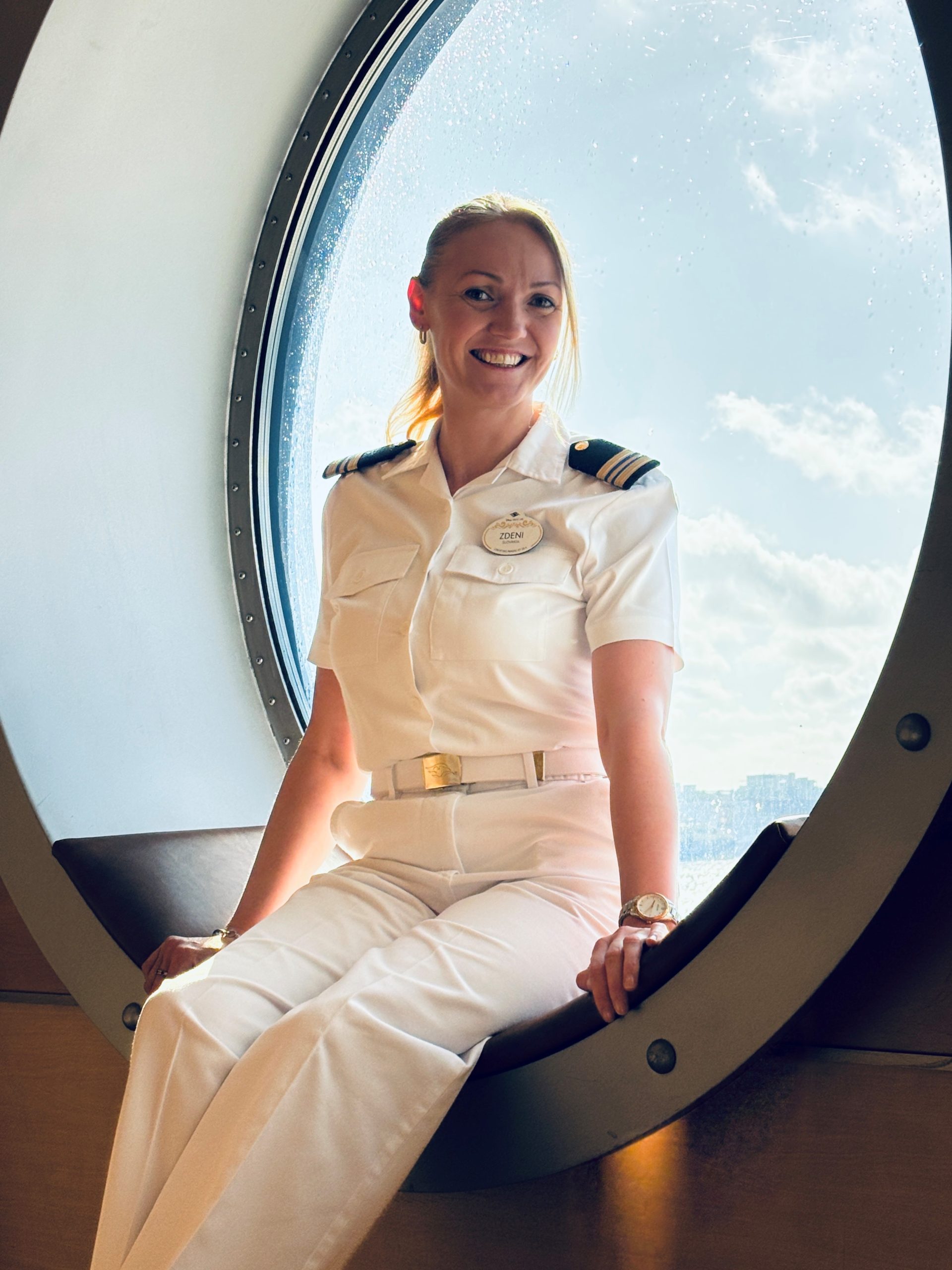 Disney Cruise Line Human Resources Manager Zdeni sits beside a large porthole window aboard a Disney cruise ship.
