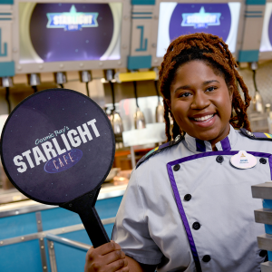 Disney college program food and beverage female cast member in uniform smiling and holding up a paddle showing the Starlight Cafe logo.