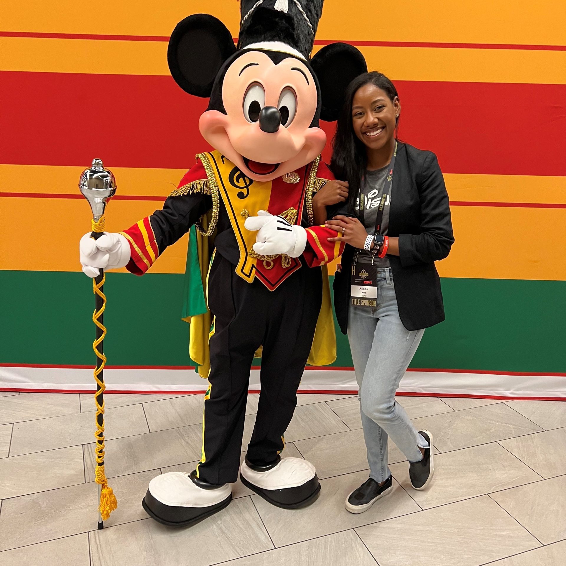 Photo of employee posing with Drum Major Mickey Mouse in front of a yellow, red and green striped backdrop.