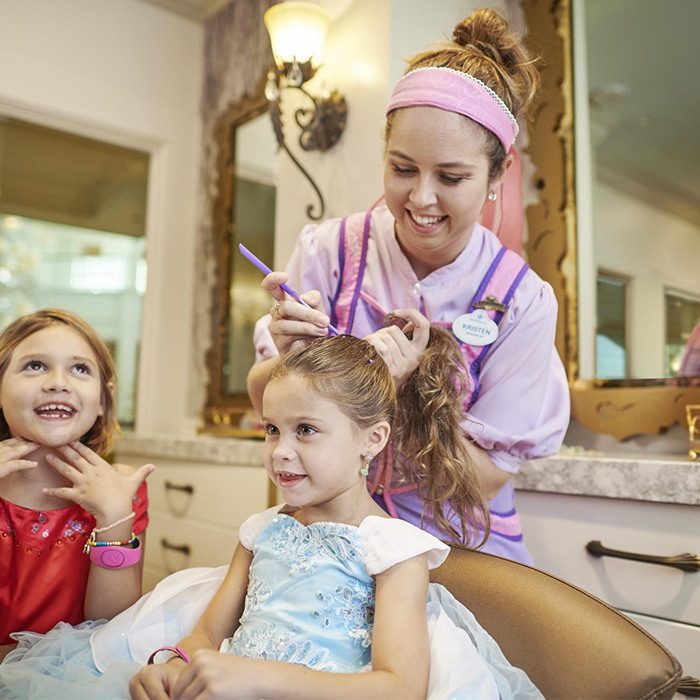 Two kids sitting in salon chairs smiling while a disney college program cast member styles their hair at the Walt Disney World Bibbidy Bobbidy Boutique