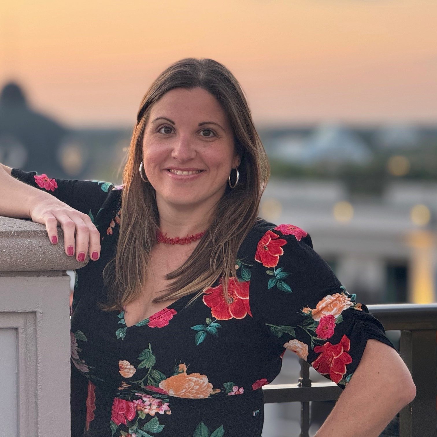 Photo of employee in a floral top with arm propped on a ledge in front of a sunset.