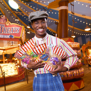 Disney college program retail and sales male cast member wearing a hat and glasses holding bags of popcorn smiling