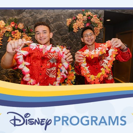 Two people in red floral shirts holding colorful leis behind a counter decorated with tropical flowers.